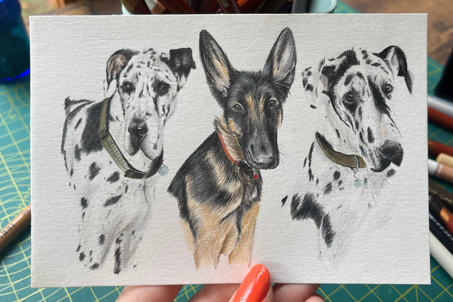 Hand holding a small sheet of paper with a drawing of a dalmatian dog, surrounded by art supplies on a table.